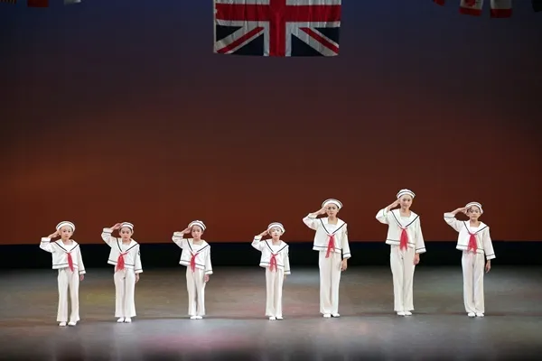 2019年の公演。万国旗を背に、セーラー衣装の子ども7名が横一列で敬礼する群舞